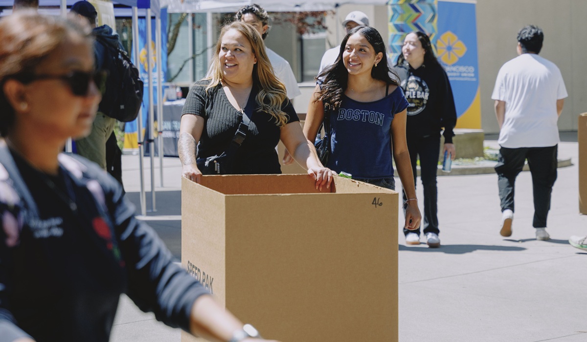 A woman helps a younger woman push a box across campus