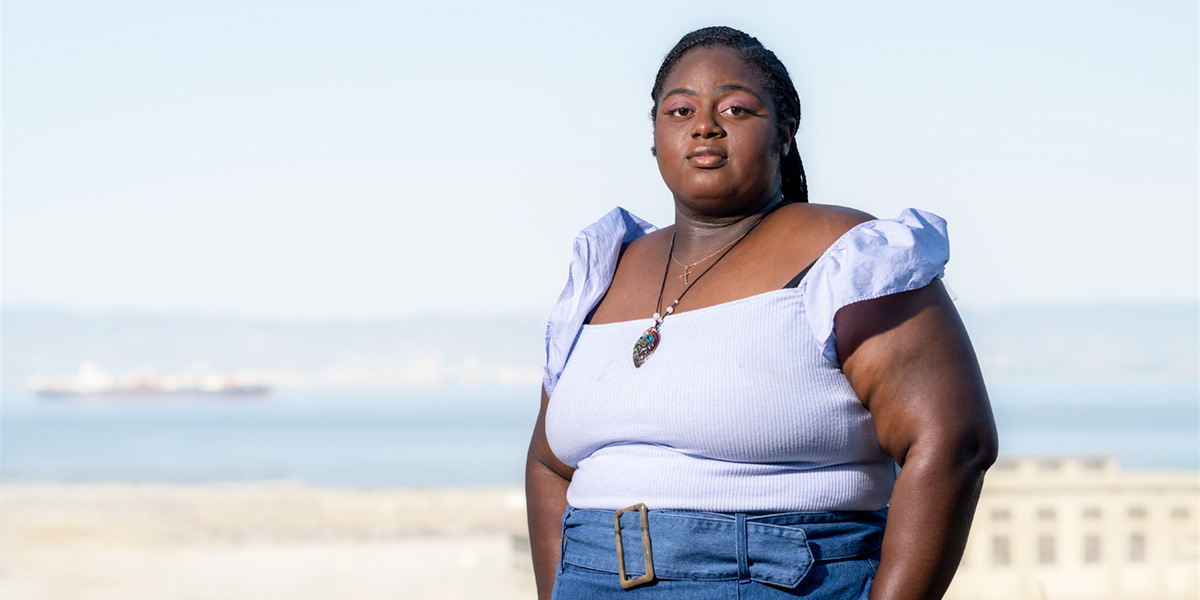 Chinomnso Okorie stands with her hands in her pants pockets on a hilltop in San Francisco on a sunny day with the Hunters Point Naval Shipyard, the San Francisco Bay and the East Bay hills in the background