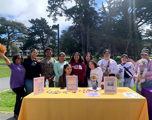 Members of the Peer2Peer Mentorship Collective, with one of them holding a dog, pose for a picture while tabling on the Quad on a partly cloudy day