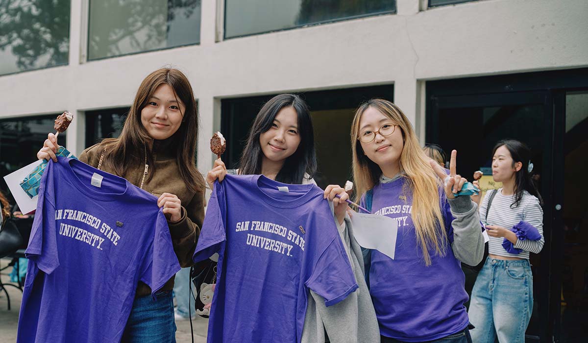 Three people hold up purple San Francisco State University T-shirts while standing and eating ice cream