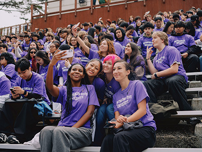 A student takes a selfie while seated with hundreds of other students in Cox Stadium wearing purple SFSU T-shirts