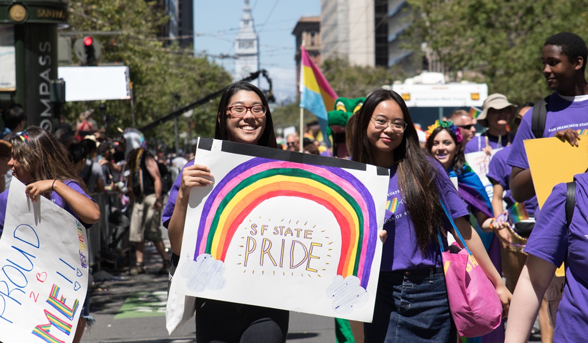 Two students marching in a parade stand in front of a hand-made sign saying &quot;SF State Pride&quot;