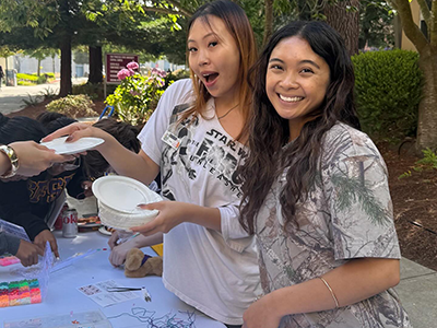 Two students smile while handing out art supplies and standing at a table on a sunny day at SFSU