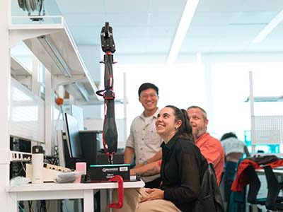 Three engineering students looking up at a robotic arm