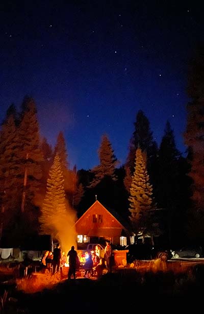 Starry night sky with trees and cabin light up with the glow from a campfire