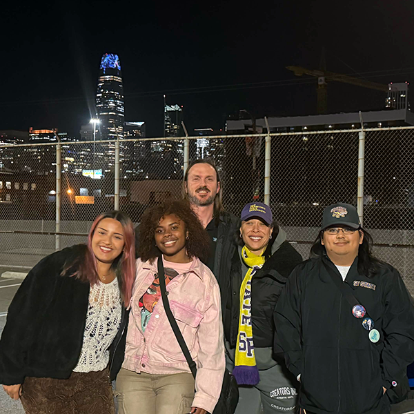 Graham Carpenter and four students pose for a photo while standing outdoors at night with their artwork featured on the Salesforce Tower and a chain-link fence in the background