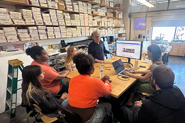 Artists Jim Campbell, Emma Strebel and three students are seated at a table in Campbell's studio and looking at a computer monitor