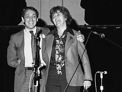 Harvey Milk places his arm on Sally Gearhart's shoulder while they stand and smile on stage at a public event