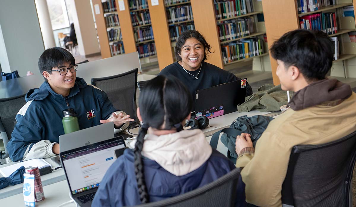 Four students sitting around a table in the library, chatting and using laptops