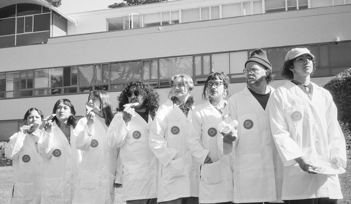 Black and white photo of eight SFSU students in lab coats standing on the Quad with four of them each eating a slice of pizza