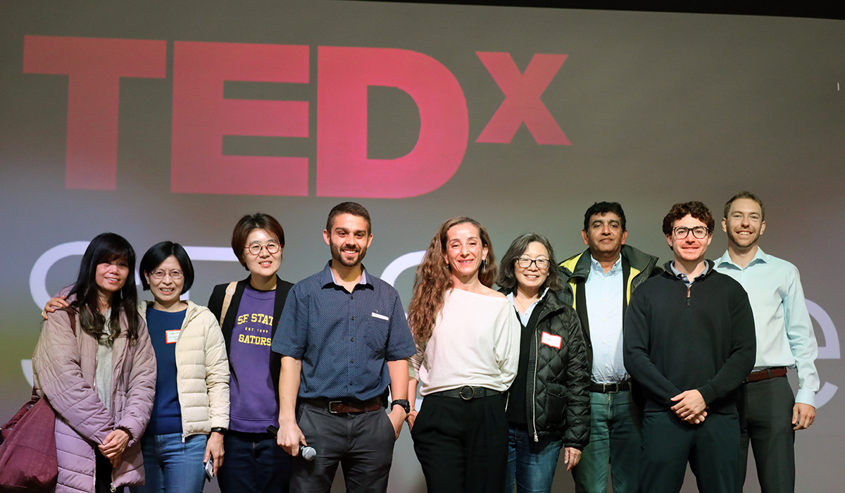 Nine students, faculty and administrators stand on stage in front of a screen projecting the TEDx logo