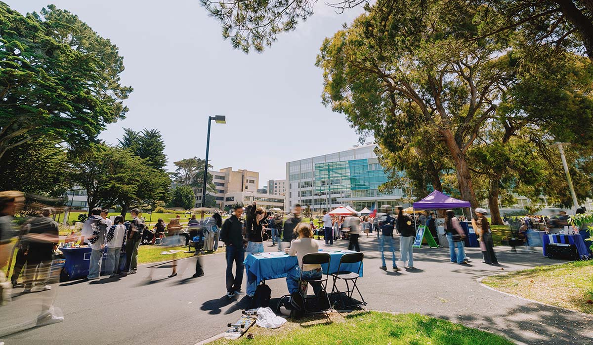 People walk across the Quad among tables on a sunny day