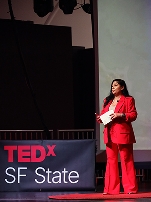 Janet Wright stands next to a table with a tablecloth showing the text "TEDx SF State" while giving her talk