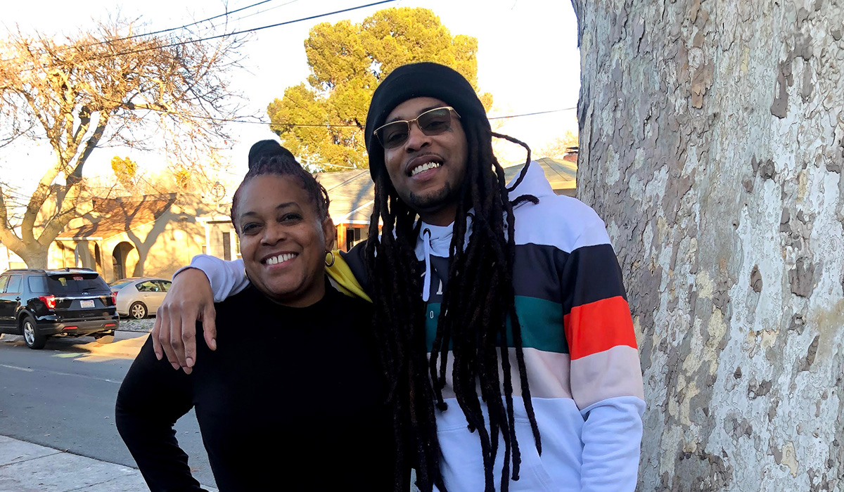 Marcus Angelo Bryant stands to the right of his mother, Theresa Thompson, with his right arm around her shoulders while both smile and stand next to a tree on a sunny day