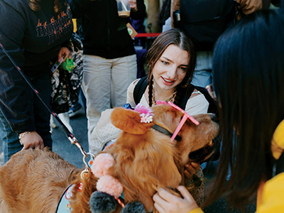 Two people crouch down to pet a dog wearing pink sunglasses at an outdoor therapy animal event at SFSU