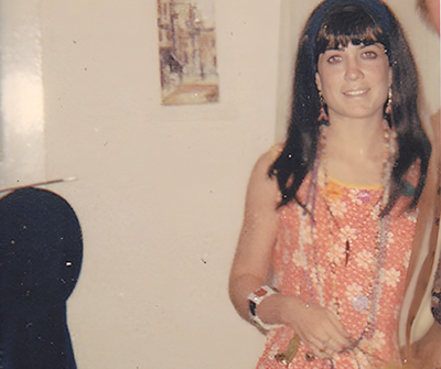 A photo from the 1960s shows Linda Yelnick standing indoors in front of a chair wearing a dress, beaded necklaces and a beaded bracelet while holding a yellow bag and a floral-designed bag