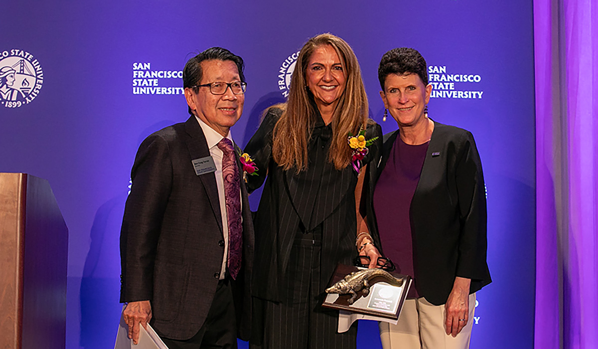 Three dressed up people pose together in front of a purple backdrop that says SFSU. 
