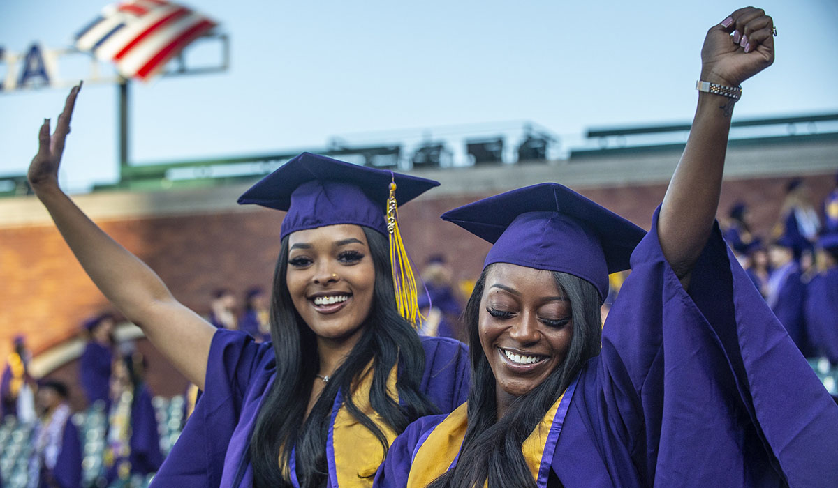 two people with long hair dressed in purple and gold reglia raise their arms in celebration