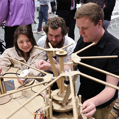 three students kneeling to look at the wooden engineering device