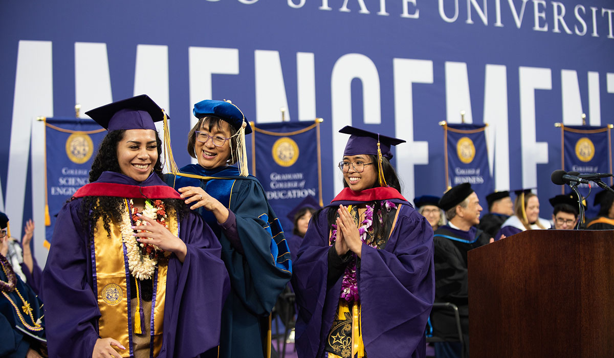 A student dressed in regalia gets hooded by a dean as another students watched proudly 