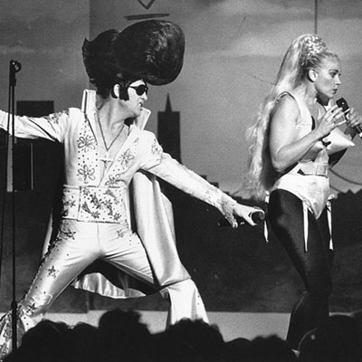 Leslie impersonates Madonna while standing next to a person impesonating Elvis and wearing an exagerated pompadour