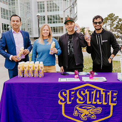 Four people stand behind a table in the Quad with water bottles on the table. 