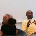 Delroy Lindo cups his hands together while seated in front of a whiteboard in a classroom wearing eyeglasses and a yellow sweater