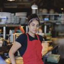 A Person in a red apron poses for a photo while standing inside a commercial kitchen