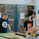 Lecturer Kevin Chen observes two students measuring small blocks of wood in the Wood Shop in the Fine Arts building