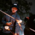 At an evening bonfire, a person lights a marshmallow on a stick while others watch 