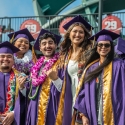 Graduates dressed in regalia stand together in the bleachers at Oracle Park