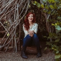 Alie Ward sitting and smiling by a tree hut