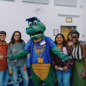 Seven people smile while posing for a photo with the Alli Gator mascot in the middle pointing their fingers horizontally and with one of the other people flashing a peace sign