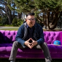 LeRoid David smiles, clasps his fingers together and looks toward his left while sitting on a purple couch on the Quad next to a blue baseball cap on a sunny day 