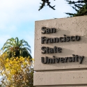 The San Francisco State University pylon sign surrounded by trees and next to a stoplight and a bicyclist road sign on a mostly sunny day
