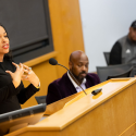 Dawn-Elissa Fischer uses hand gestures while speaking at a lectern dressed in black while other people seated next to hear watch