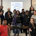 The SF State Forensics team poses for a picture in a classroom with Kivraj “Ki” Singh at top left holding a trophy from the American Forensics Association National Speech Tournament