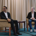 Laxman Narasimhan and Chris Larsen look to their right while seated on a stage between a table and in front of a curtain