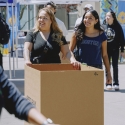 A woman helps a younger woman push a box across campus