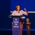 Nancy Pelosi gestures with her left hand while speaking at a lectern with the text San Francisco State University