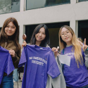 Three people hold up purple San Francisco State University T-shirts while standing and eating ice cream