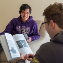 A person seated looks at a book while another person seated across from them smiles while wearing a purple SFSU hoodie