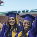two people with long hair dressed in purple and gold reglia raise their arms in celebration