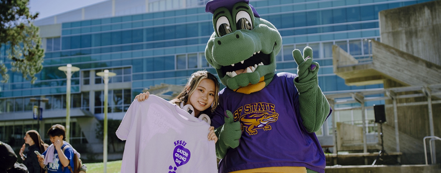Student holding a lavendar Gator Giving Day shirt poses next to Alli Gator