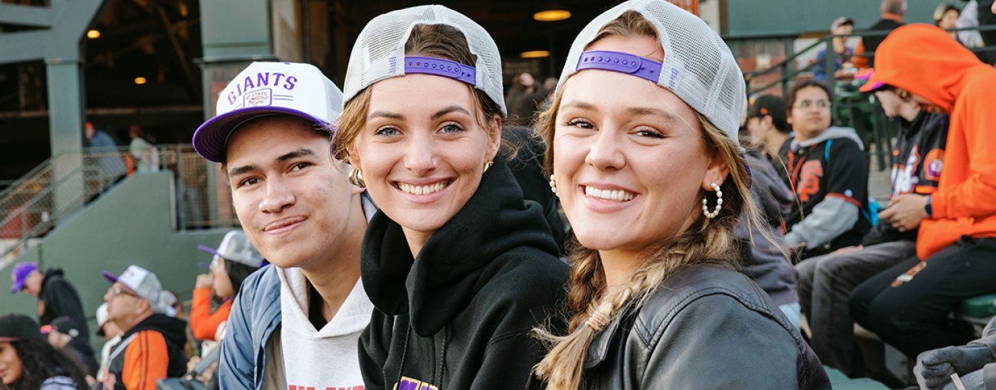 Three people smile while seated and wearing co-branded SFSU-San Francisco Giants baseball caps at Oracle Park