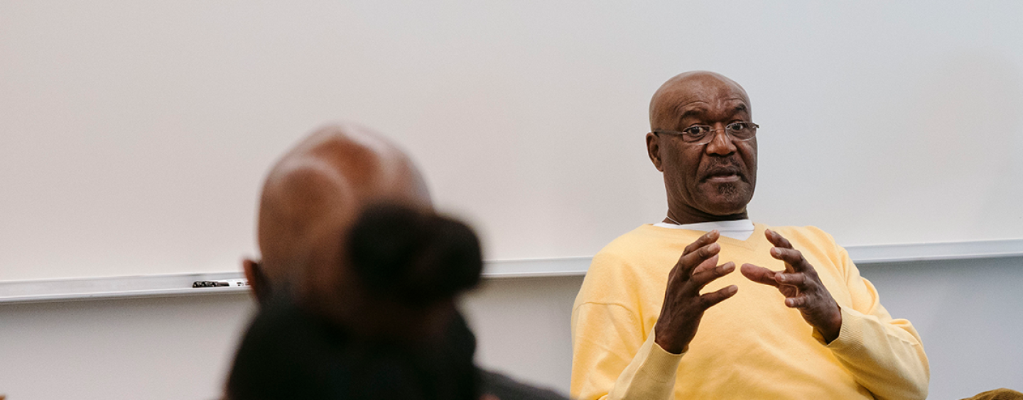 Delroy Lindo cups his hands together while seated in front of a whiteboard in a classroom wearing eyeglasses and a yellow sweater