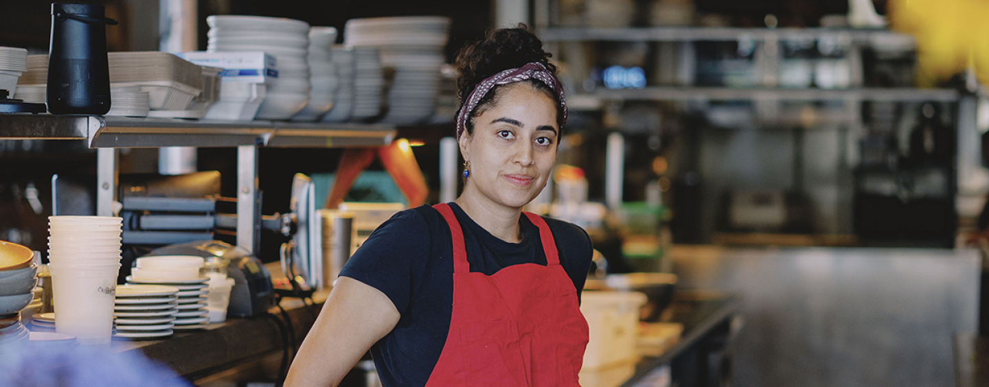 A Person in a red apron poses for a photo while standing inside a commercial kitchen
