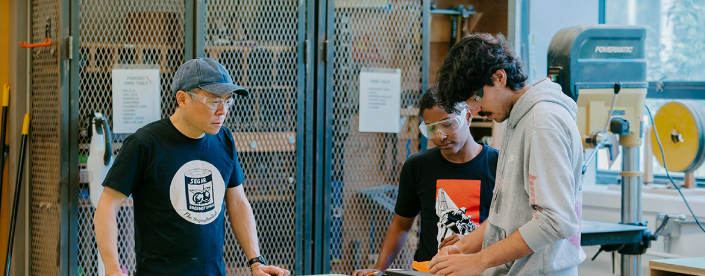 Lecturer Kevin Chen observes two students measuring small blocks of wood in the Wood Shop in the Fine Arts building