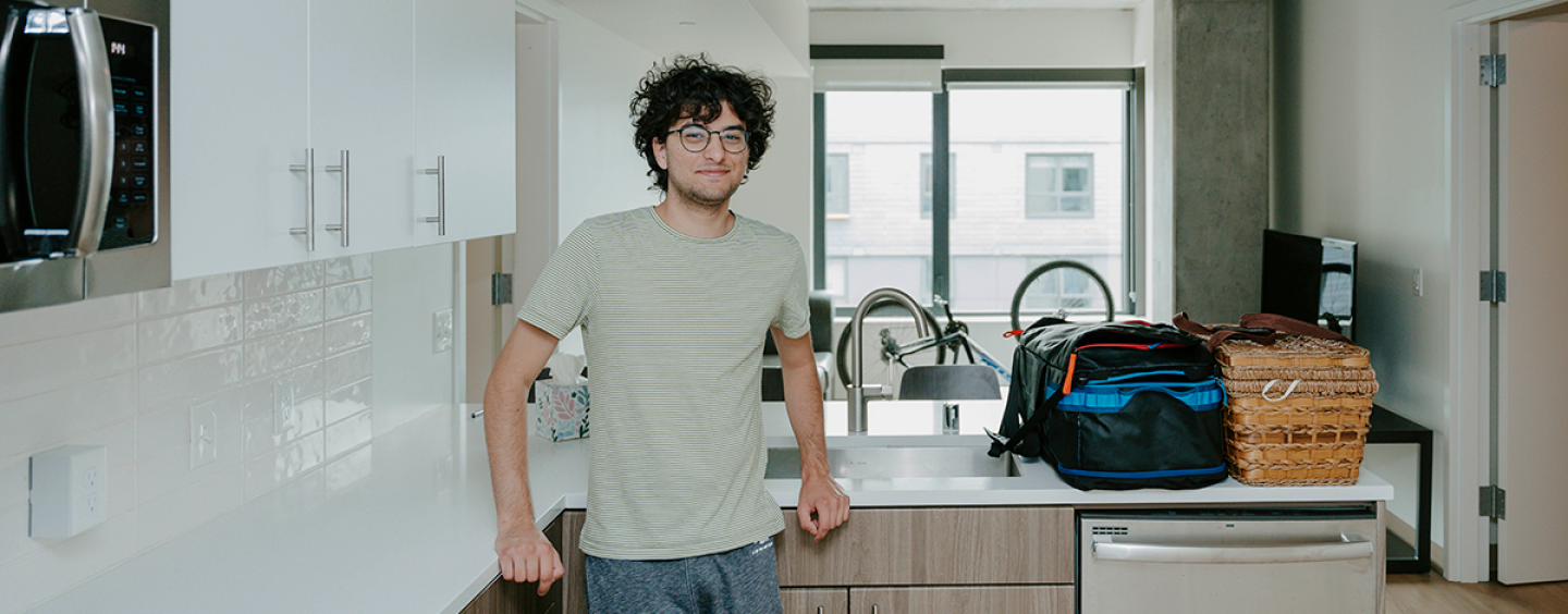 A student leans against the counter in his kitchenette in a residence hall with bags, a bicycle and a window behind him