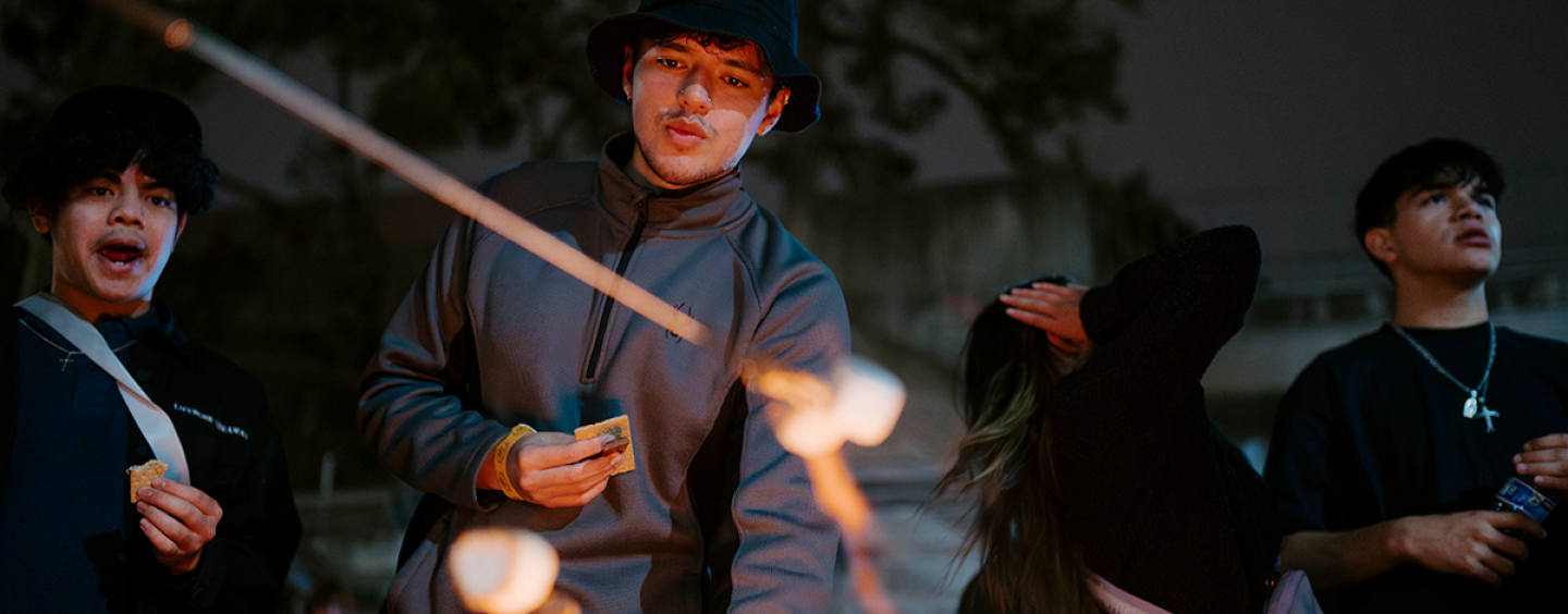 At an evening bonfire, a person lights a marshmallow on a stick while others watch